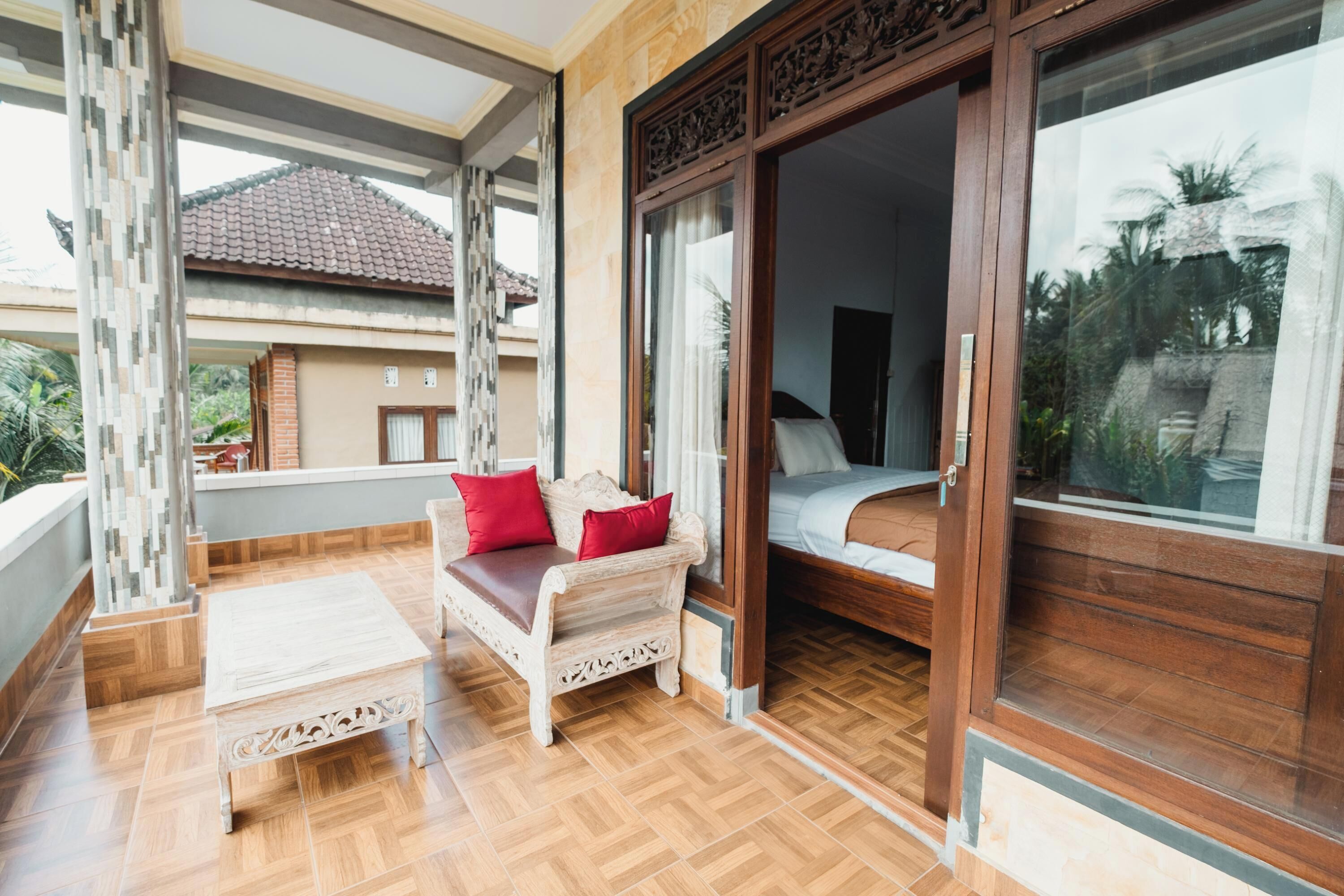 A balcony with a wooden bench and a small table, a sliding glass door leading to a bedroom, and a view of a building with a tiled roof.