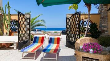 A beachside patio with two striped lounge chairs, a wicker table with flowers, and a view of the ocean.