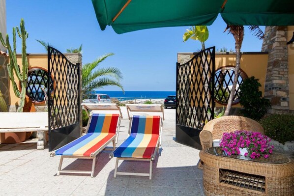 A beachside patio with two striped lounge chairs, a wicker table with flowers, and a view of the ocean. - Lovely 1-bed Villa in Balestrate (Balestrate)
