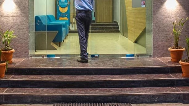 Un hombre está de pie frente a la entrada de un edificio con puerta de vidrio, un área de descanso azul en su interior y macetas a ambos lados de la entrada.