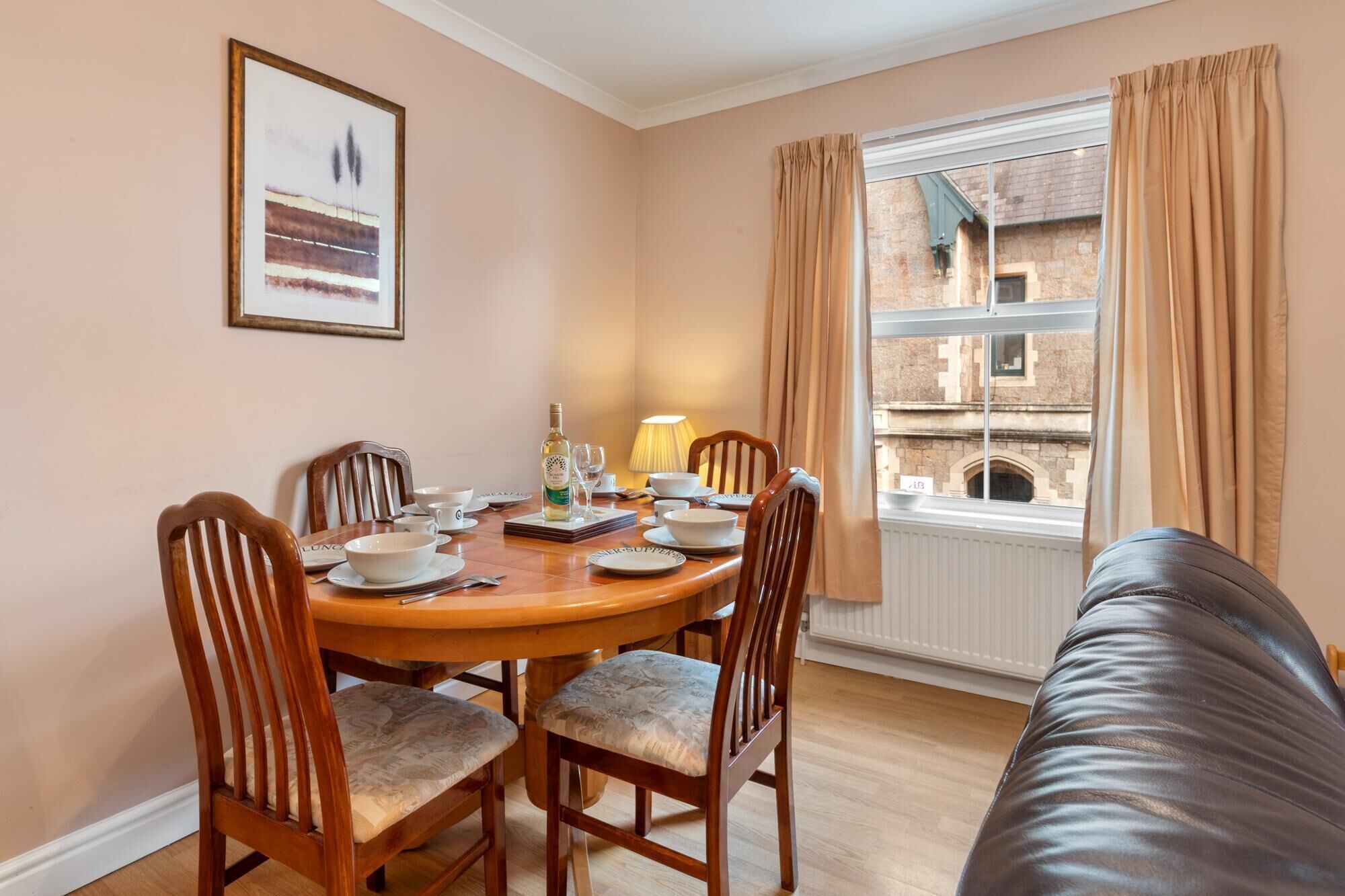 A dining area with a wooden table set for four, surrounded by chairs with cushioned seats. A bottle of wine and a lamp are on the table. A framed picture hangs on the wall, and a window with curtains is visible.