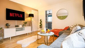 Ein modernes Wohnzimmer mit einem Flachbildfernseher, der 'NETFLIX' zeigt, einem weißen Sofa, einem runden Spiegel und einem kleinen runden Tisch mit Früchten und Getränken.