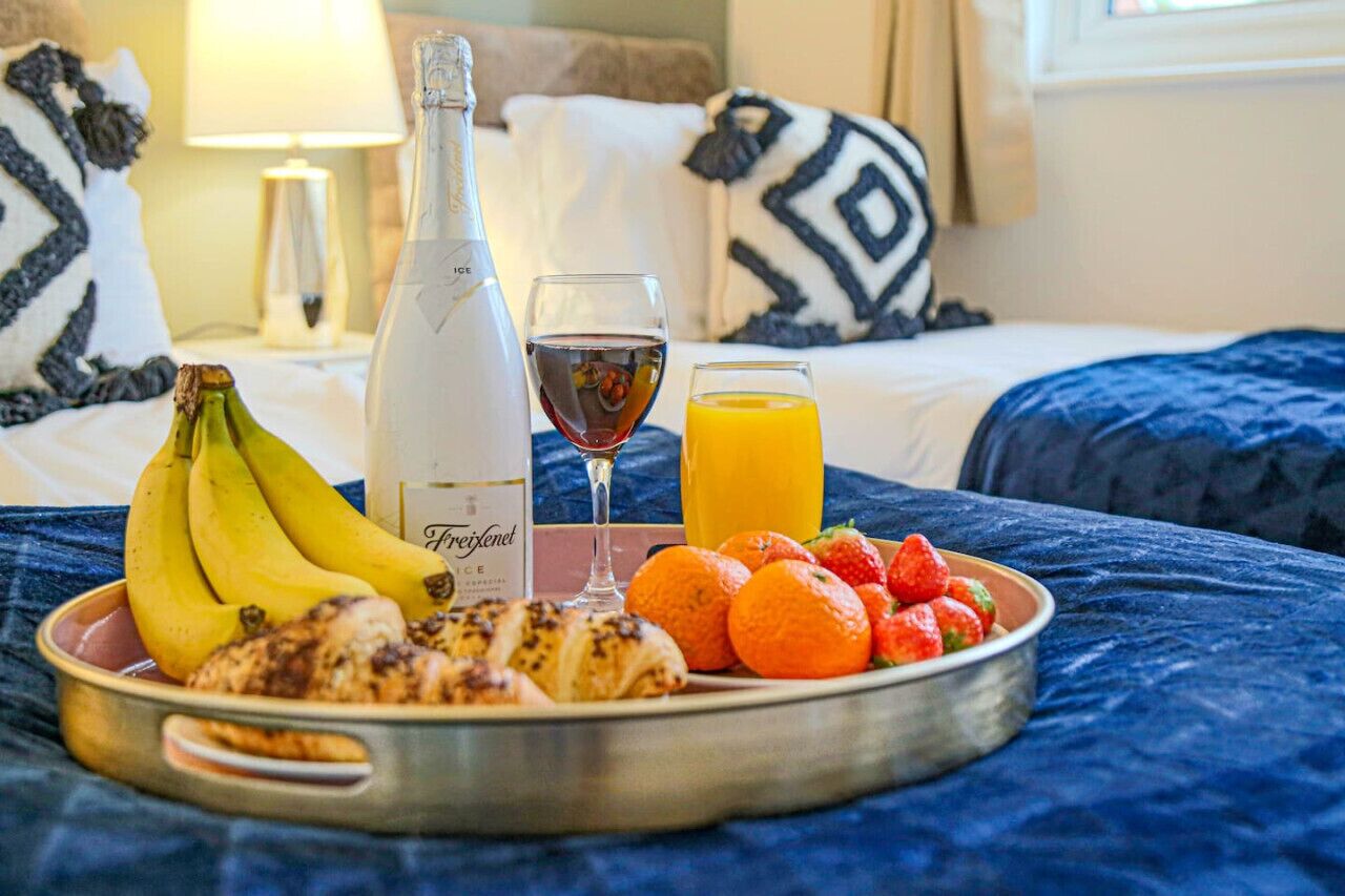 A breakfast tray with bananas, croissants, strawberries, oranges, a bottle of wine, and a glass of orange juice on a bed with a blue blanket.
