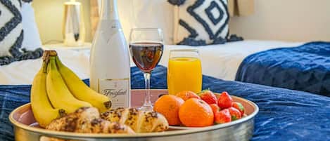 A breakfast tray with bananas, croissants, strawberries, oranges, a bottle of wine, and a glass of orange juice on a bed with a blue blanket.
