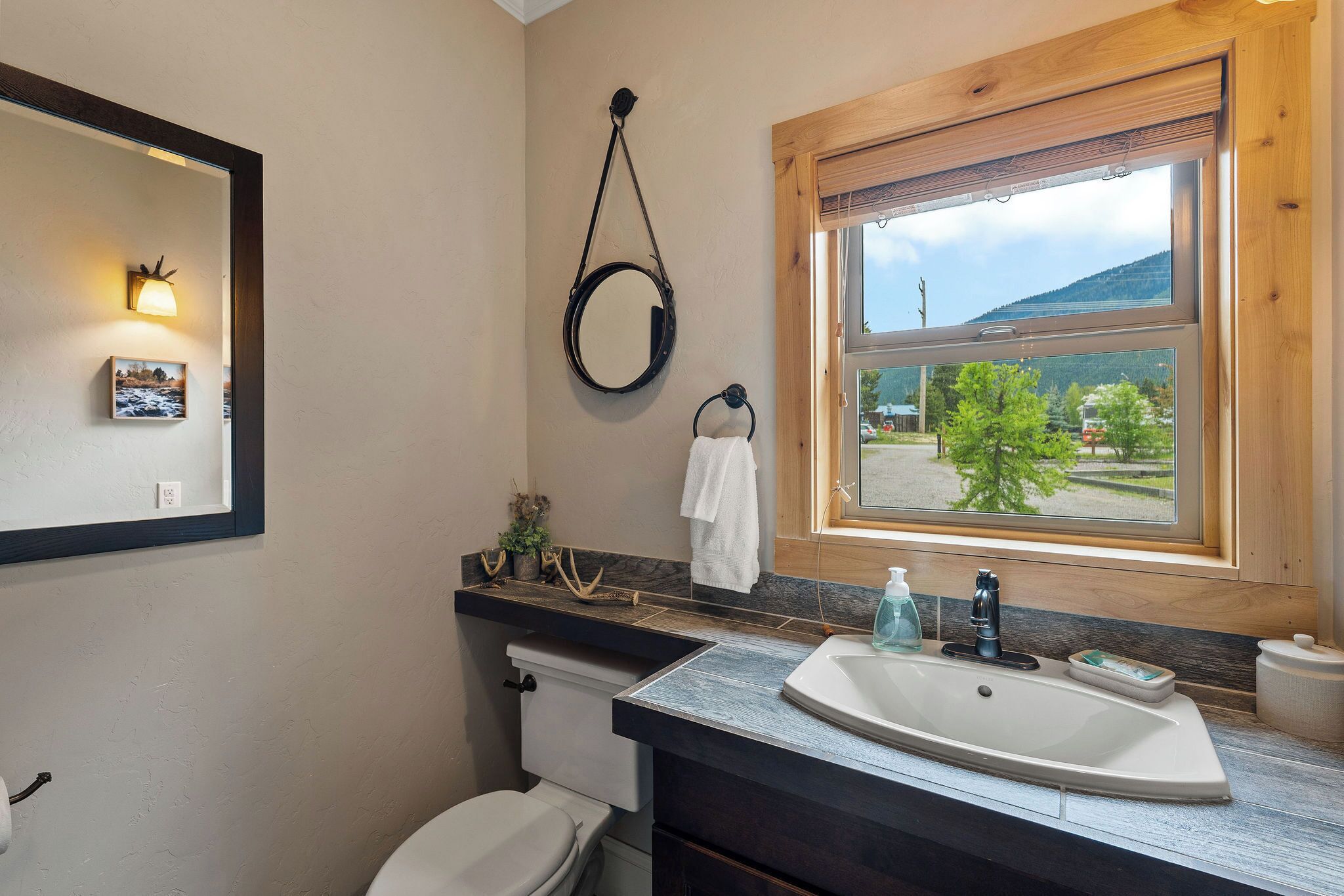 Un baño con inodoro, lavamanos, espejo y ventana con vistas a la vegetación.
