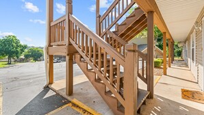 Una escalera de madera conduce a un porche cubierto, con vista despejada a la calle y follaje al fondo.