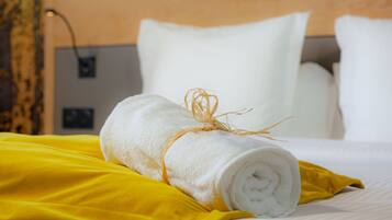Toalha branca bem dobrada, enrolada e amarrada com um barbante, colocada sobre um tecido amarelo sobre uma cama com almofadas brancas.