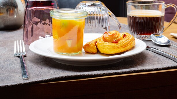 Desayuno con un croissant, un vaso de jugo de naranja y un vaso de bebida oscura sobre una mesa.
