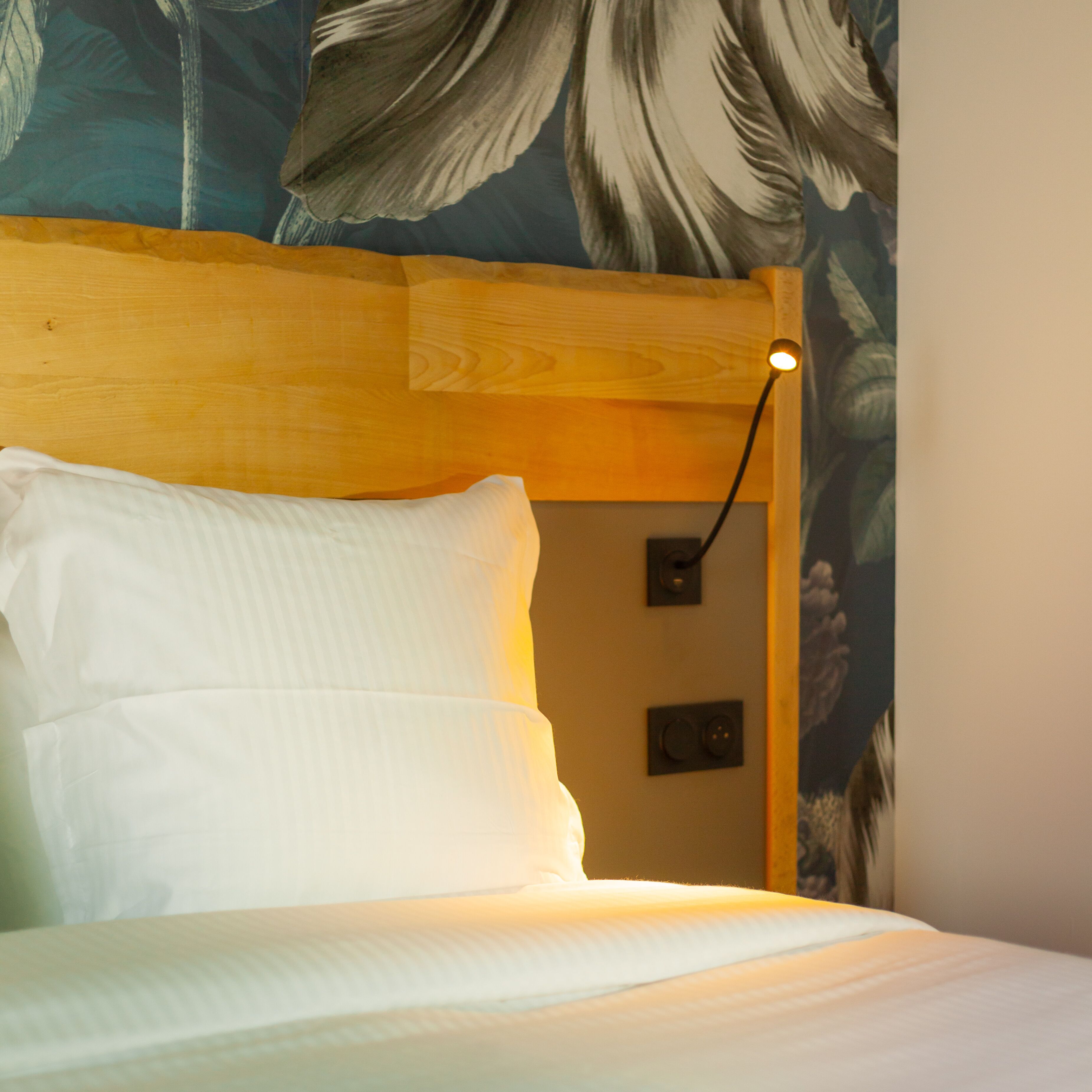 A bed with white bedding, a wooden headboard, and a wall-mounted light fixture.