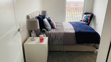 Quarto com cama, mesa de cabeceira, abajur e janela com vista para edifícios.
