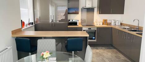 Uma cozinha moderna com ilha central, eletrodomésticos embutidos e sala de jantar com mesa de vidro e cadeiras estofadas.
