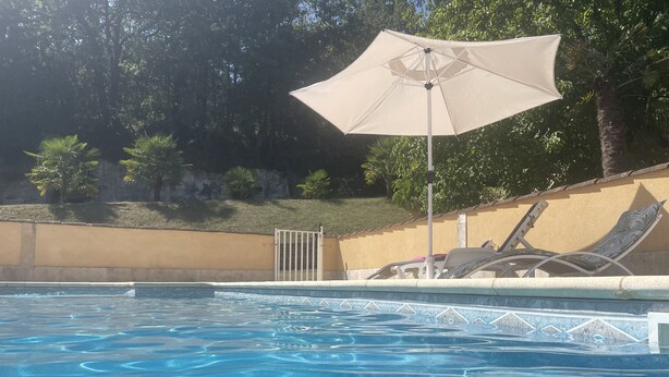 Une piscine à l’eau d’un bleu limpide, entourée d’un petit muret et d’un portail. On y trouve des chaises longues et un grand parasol qui offre de l’ombre.