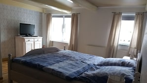 A bedroom with a bed, a television on a cabinet, a window with curtains, and a radiator. - Vacation home, North Sea, Krummhörn, Upleward <br> (Krummhörn)