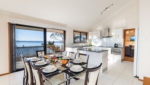 A modern kitchen with a central island, large windows with a view of the sea, and a dining area set for six. - Sea La Vie 3 Warruga St, Corlette (Corlette)