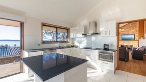 A modern kitchen with white cabinets, a black countertop, and a large window overlooking a scenic view. - Sea La Vie 3 Warruga St, Corlette (Corlette)