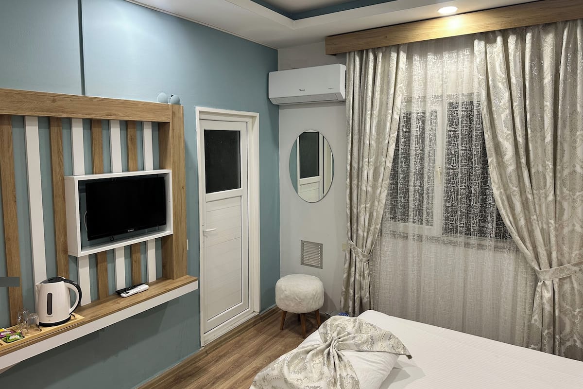 A hotel room with a TV on a wooden shelf, a door, a mirror, a window with curtains, and a bed.