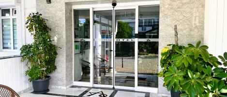 Entrada de hotel con puerta de cristal, una escalera que sube y plantas en macetas a ambos lados.