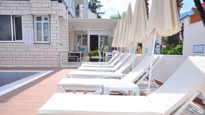 A poolside area with white lounge chairs and umbrellas, a building with a brick facade, and a clear sky. - Amsterdam Otel & Suites  (Kemer)