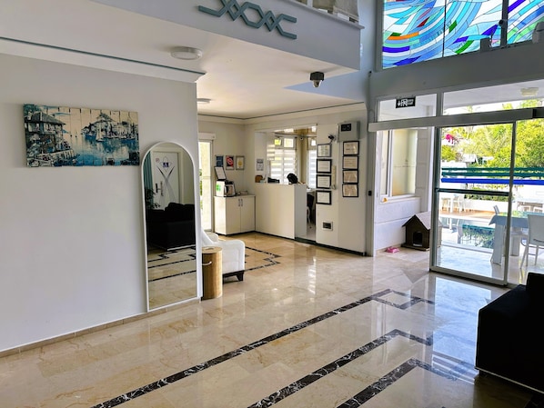 A hotel lobby with a reception desk, framed certificates, a large mirror, and a view of the outdoor area.