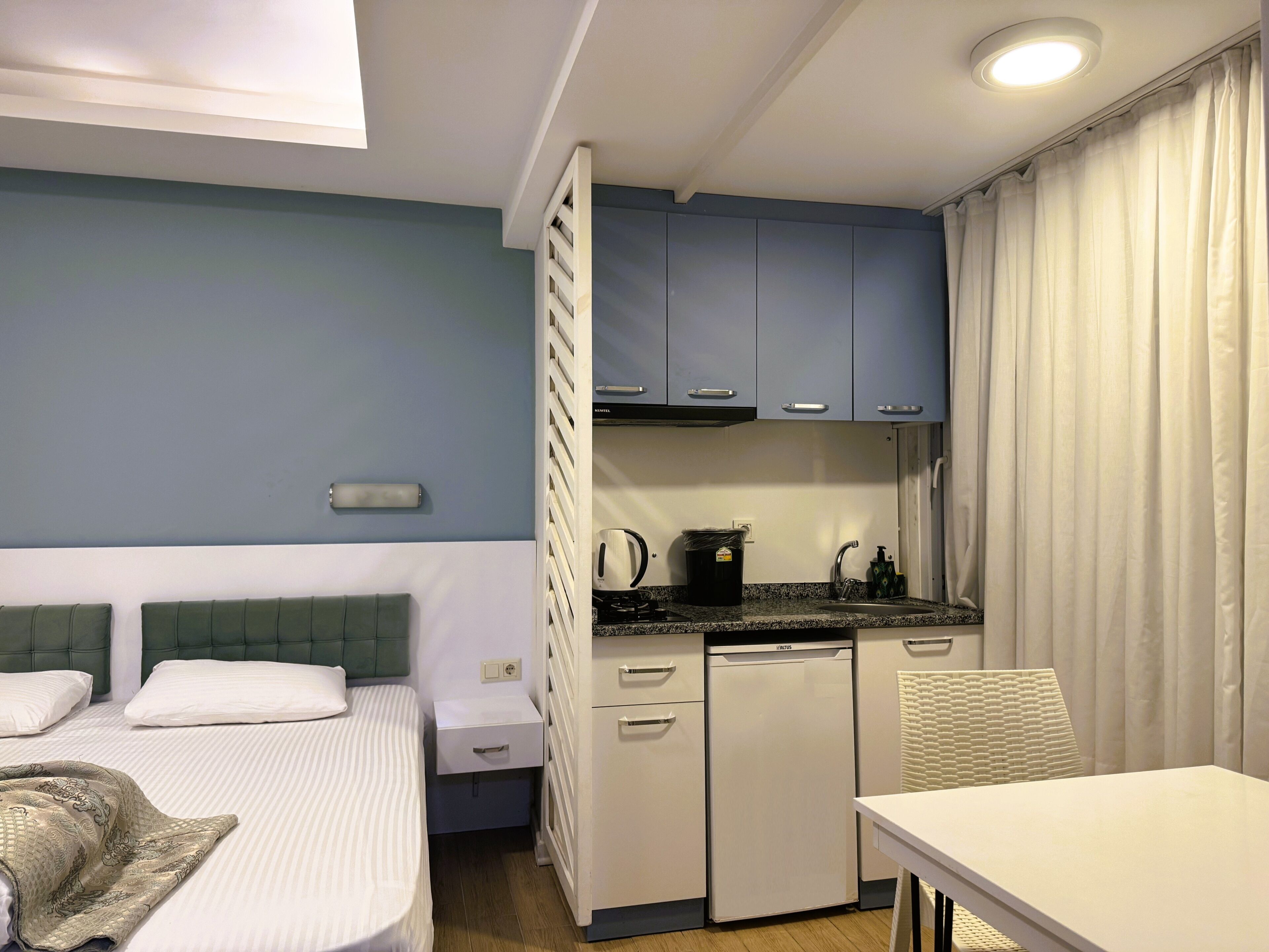 A compact hotel room with a bed, a small kitchenette, a table, and a chair.