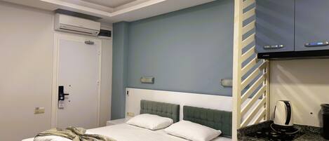 Habitación de hotel con cama, cocina integrada con estufa, puerta blanca y unidad de aire acondicionado.