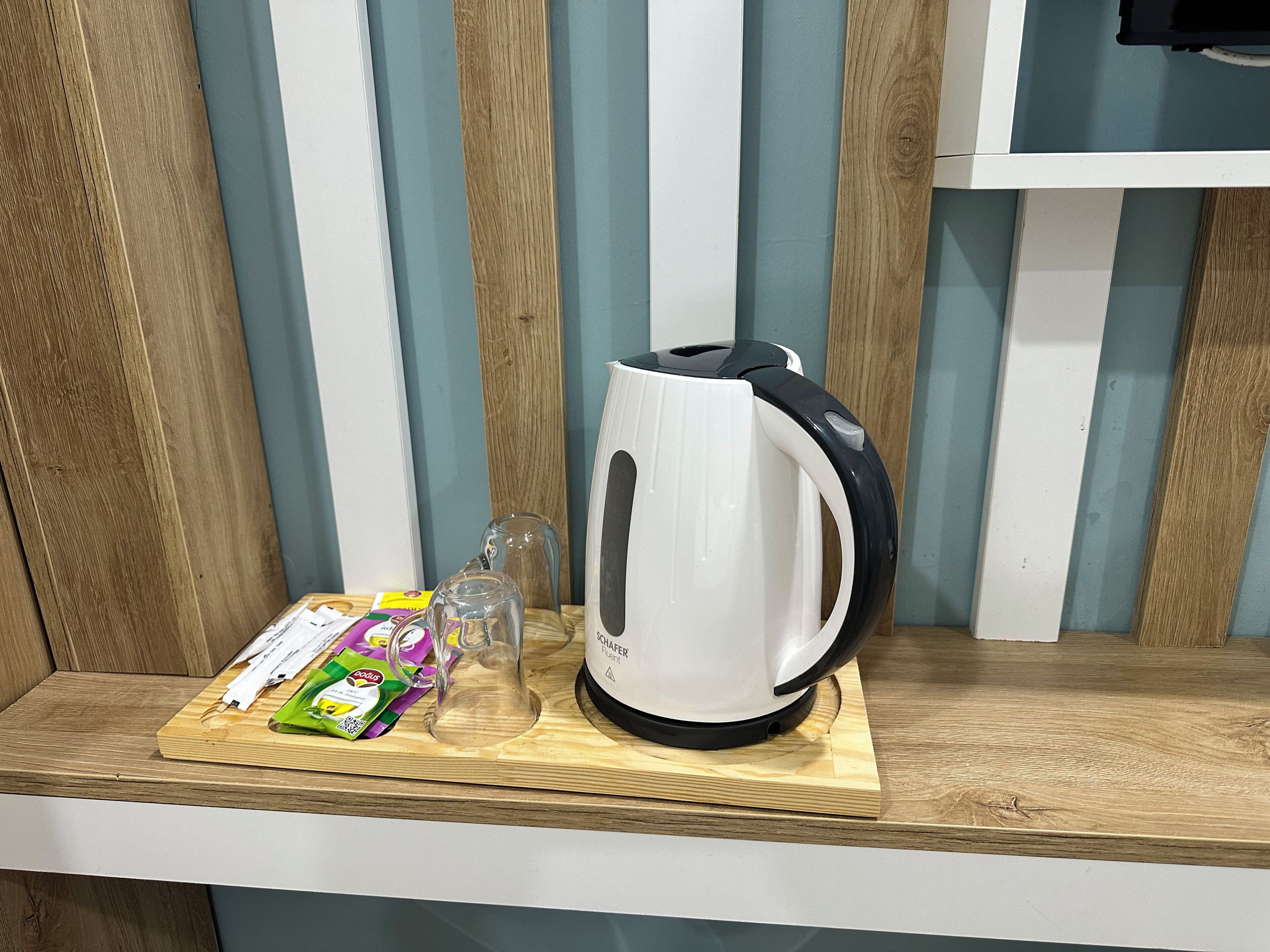 A white electric kettle on a wooden shelf with a glass pitcher and packets of tea on a cutting board.