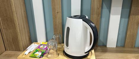 Una tetera eléctrica blanca sobre una estantería de madera, junto a una jarra de vidrio y bolsitas de té sobre una tabla de cortar.