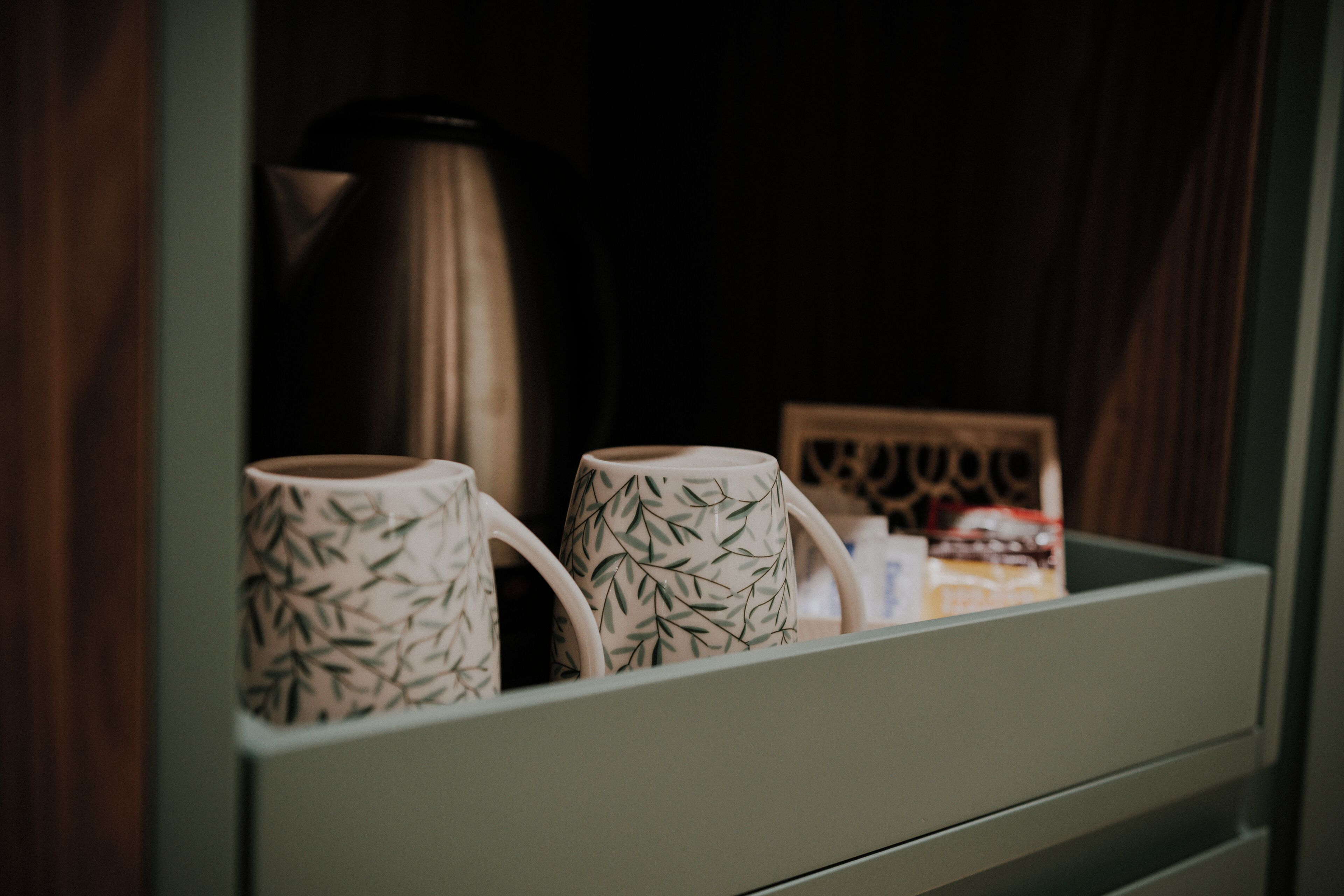 A drawer with two white mugs with a bamboo pattern, a stainless steel kettle, and a small framed picture.