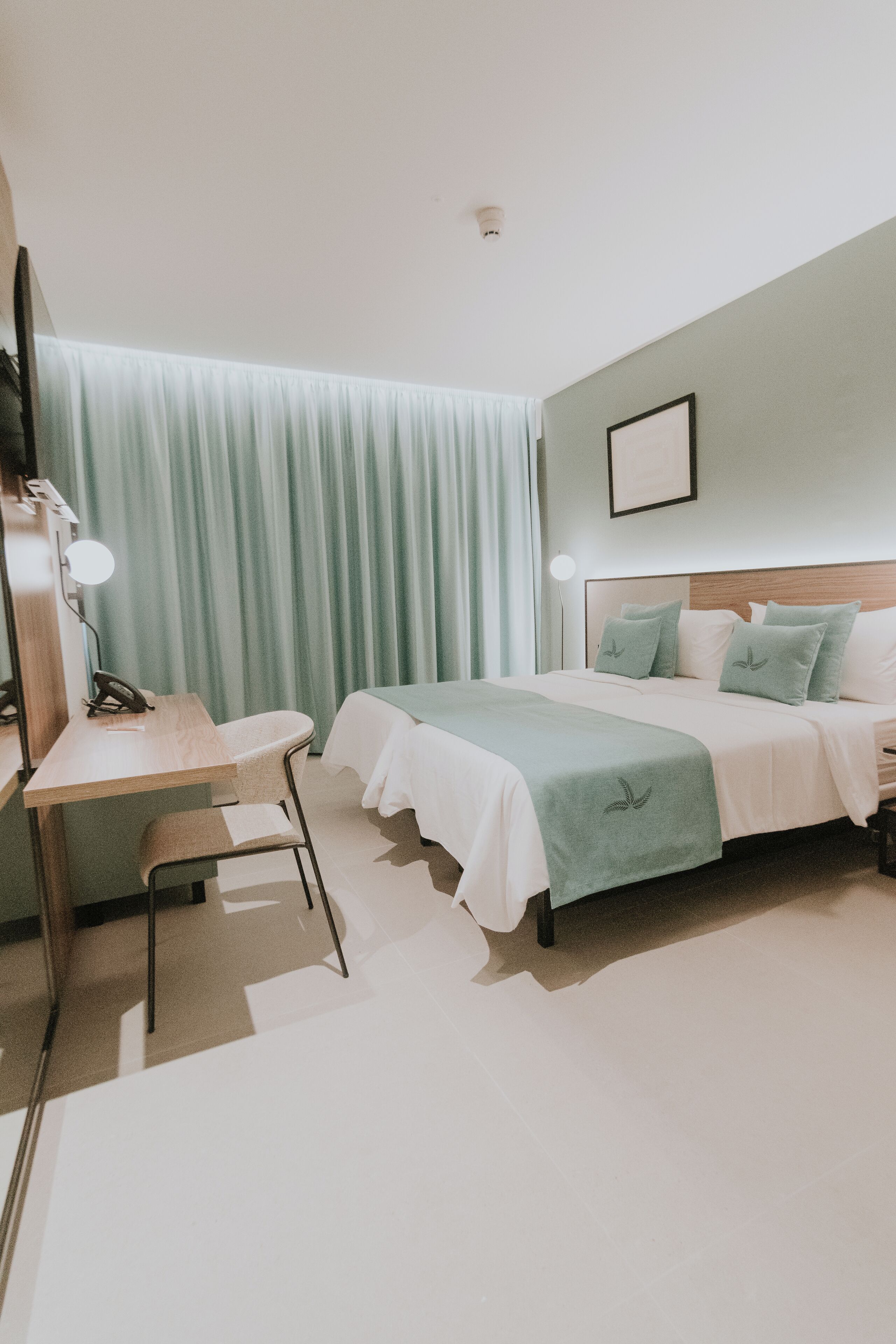 A hotel room with a bed, a desk, a chair, a lamp, and a curtain.