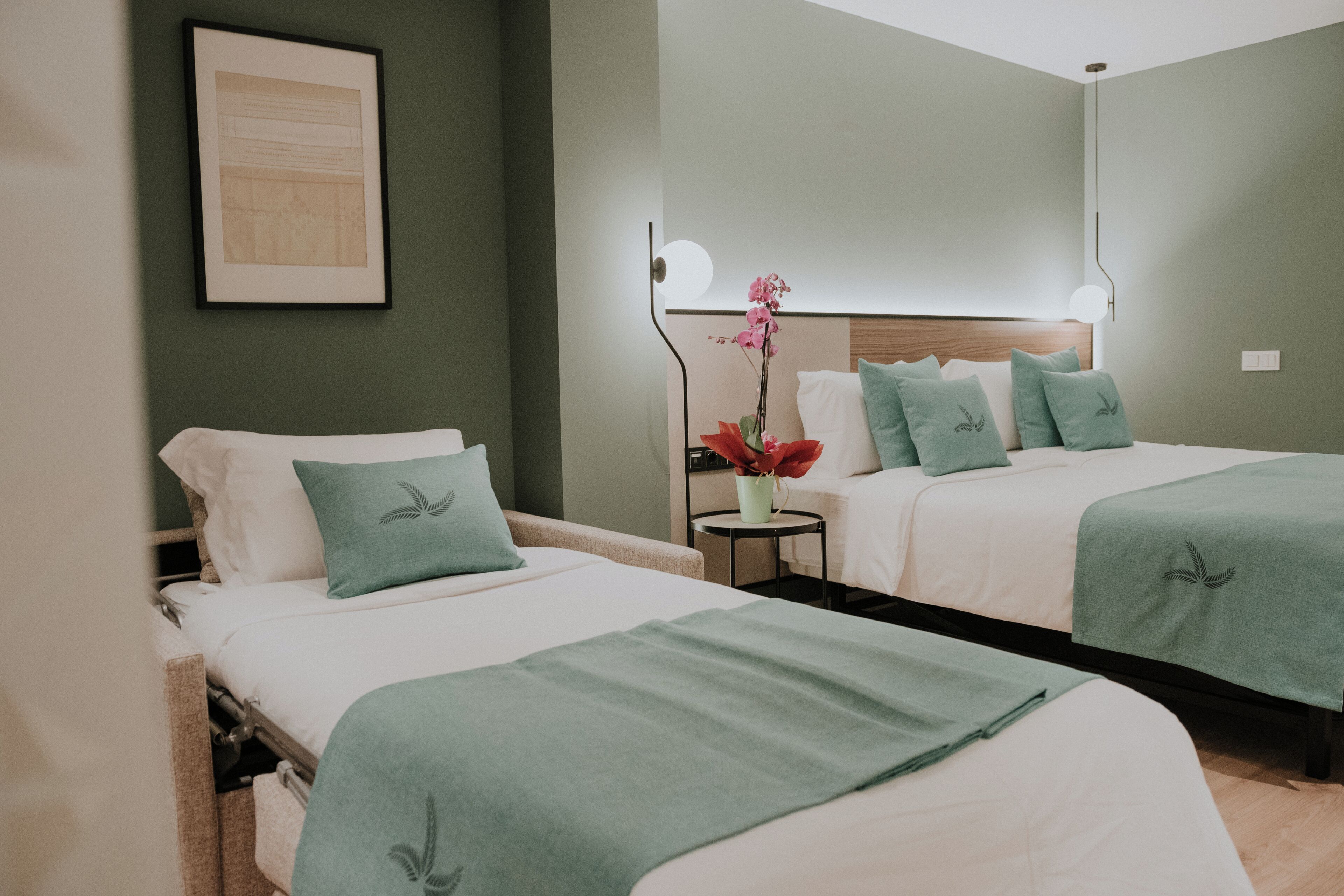 A hotel room with two beds, each with a green bedspread and matching pillows. There is a small round table with a vase of flowers between the beds.