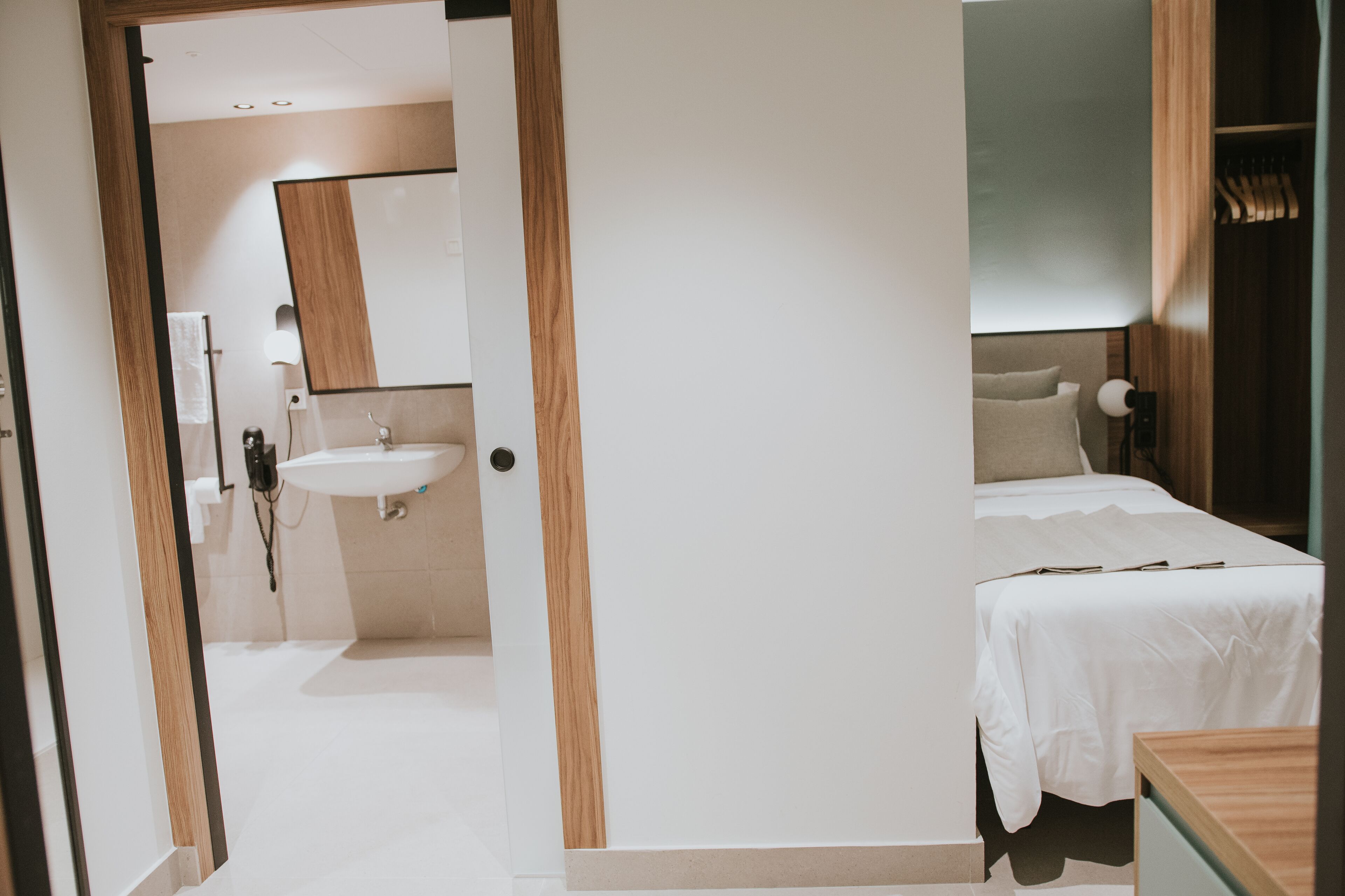 A hotel room with a wooden door, a white sink, a bed with a white bedspread, and a bedside table.
