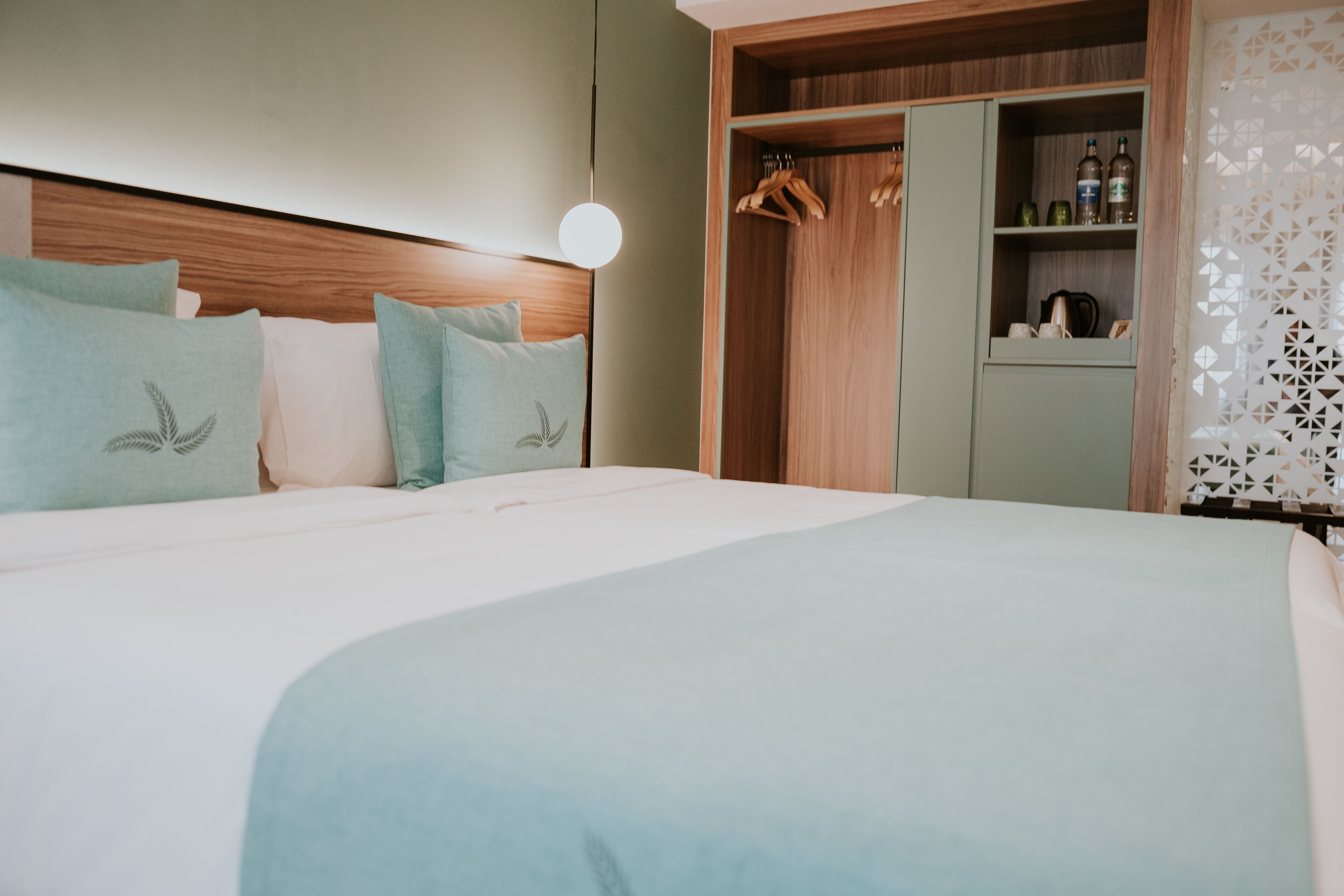 Une chambre d’hôtel avec un grand lit, une tête de lit en bois, une armoire où sont suspendus des vêtements et une étagère avec des bouteilles.
