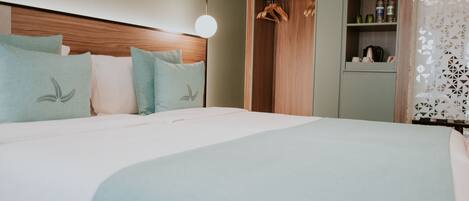 A hotel room with a large bed, a wooden headboard, a wardrobe with hanging clothes, and a shelf with bottles.
