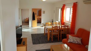 Sala de estar com sofá, mesa de centro e área de jantar com mesa e cadeiras. Há uma televisão no canto e uma janela com cortinas.