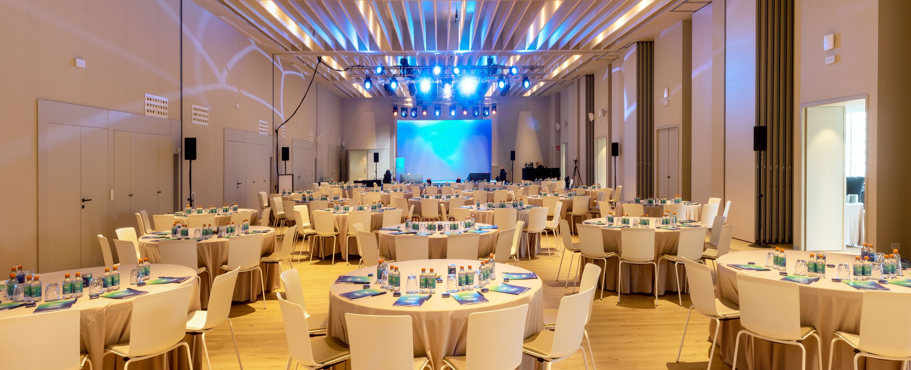 A conference room with multiple round tables, each with water bottles and name cards, and a stage with a blue-lit backdrop.