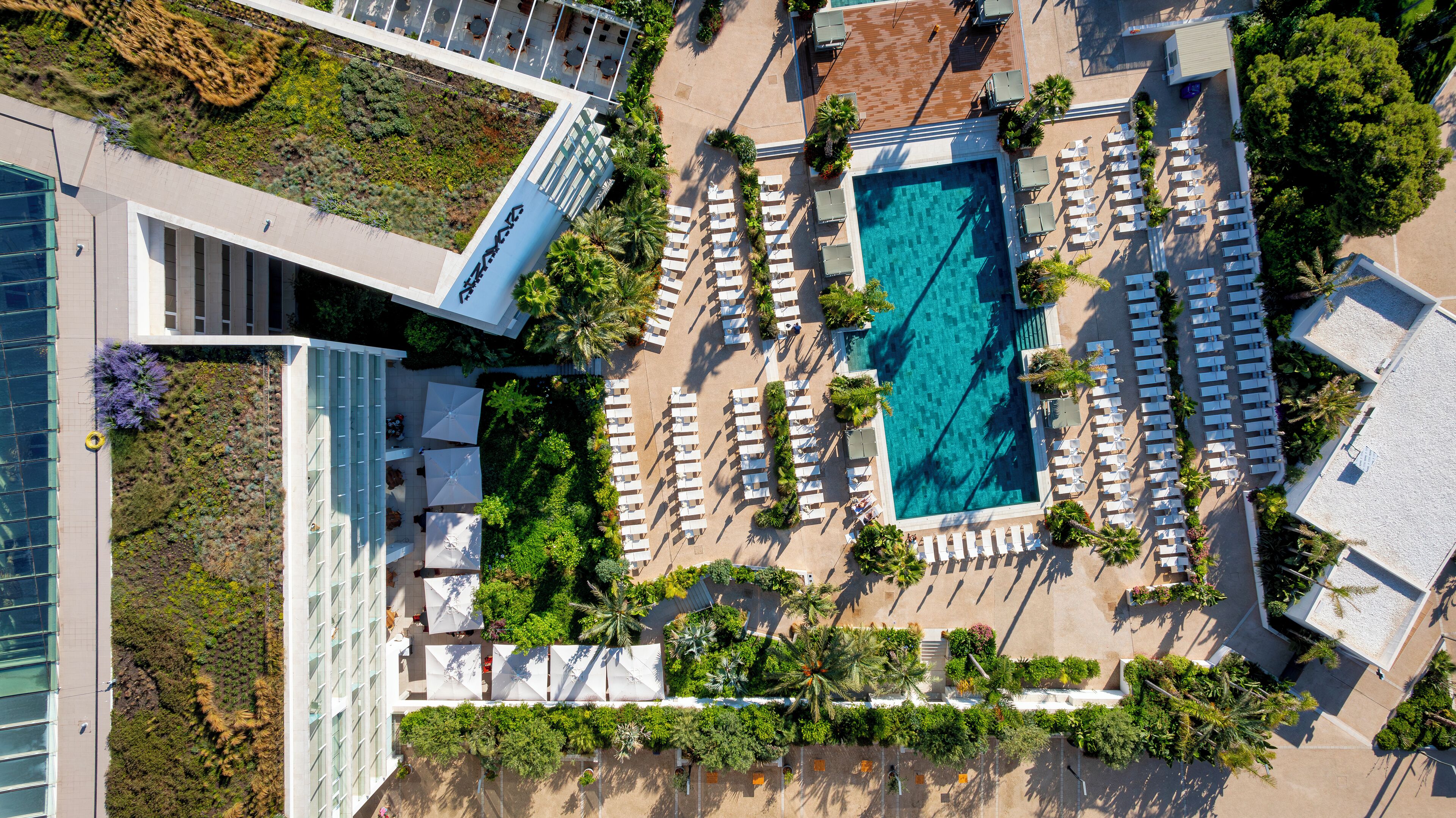 Luchtbeeld van een hotel met een zwembad, omgeven door ligbedden en parasols, een aangelegde tuin en een modern gebouw met een glazen gevel.