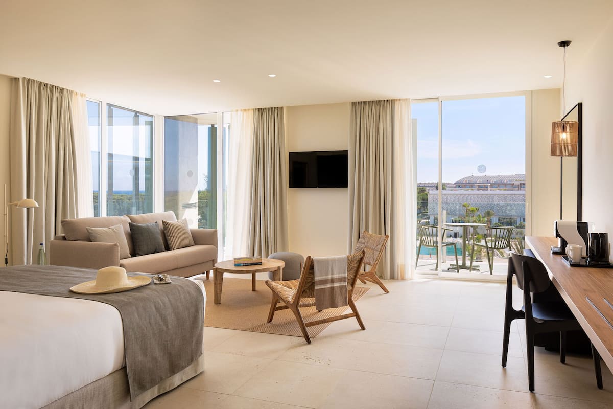Ein modernes Hotelzimmer mit einem großen Bett, einer Couch, einem Esstisch und einem Stuhl. Ein Fernseher ist an der Wand befestigt und es bietet einen Blick auf die Stadt durch große Fenster.
