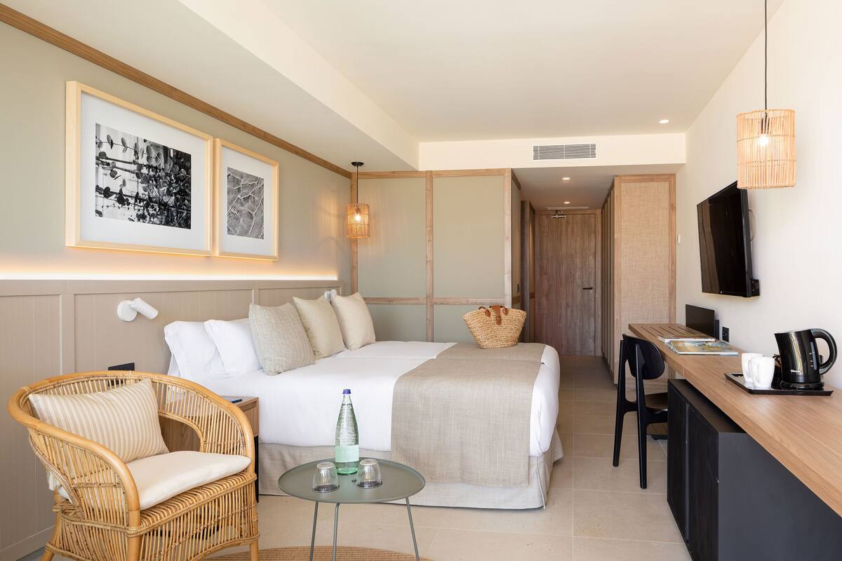 Ein modernes Hotelzimmer mit Bett, einem Korbstuhl, einem kleinen Tisch mit Gläsern und einer Flasche, einem Schreibtisch mit Kaffeemaschine und gerahmten Kunstwerken an der Wand.