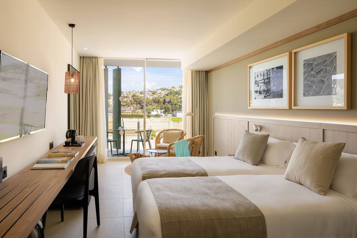 Ein Hotelzimmer mit zwei Betten, einem Schreibtisch und einem Sessel. Durch das Fenster ist die Stadt zu sehen.