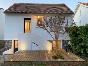 Ein modernes, zweistöckiges Haus mit Ziegeldach, Holzterrasse und einem kleinen Baum im Vorgarten.