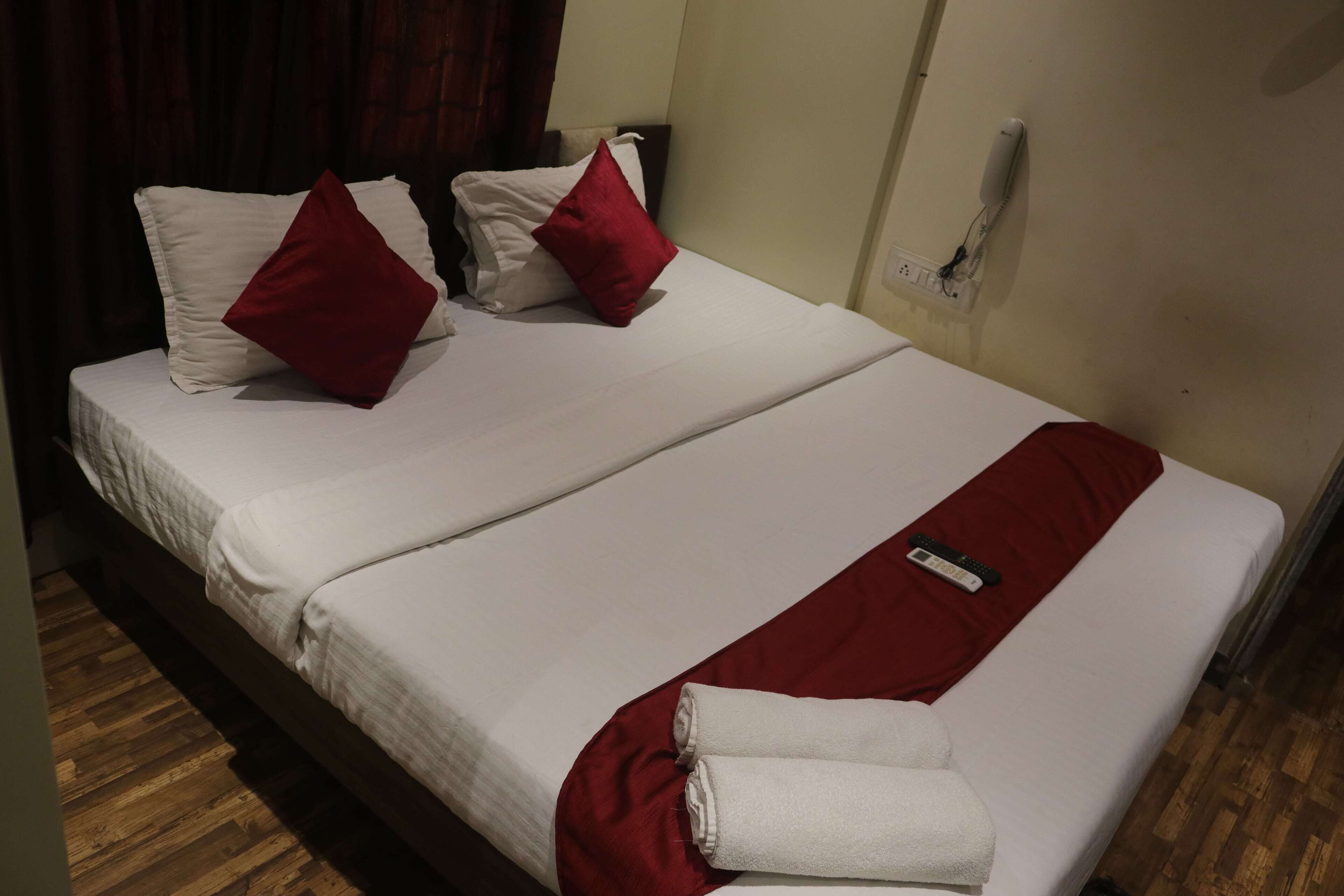 A double bed with white and red bedding, a remote control, and a rolled towel on the side.