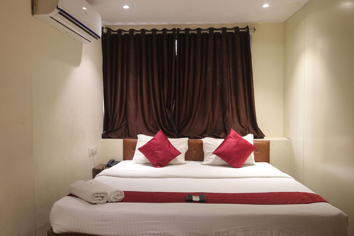 A hotel room with a bed, a red throw blanket, a phone, and a window with brown curtains.