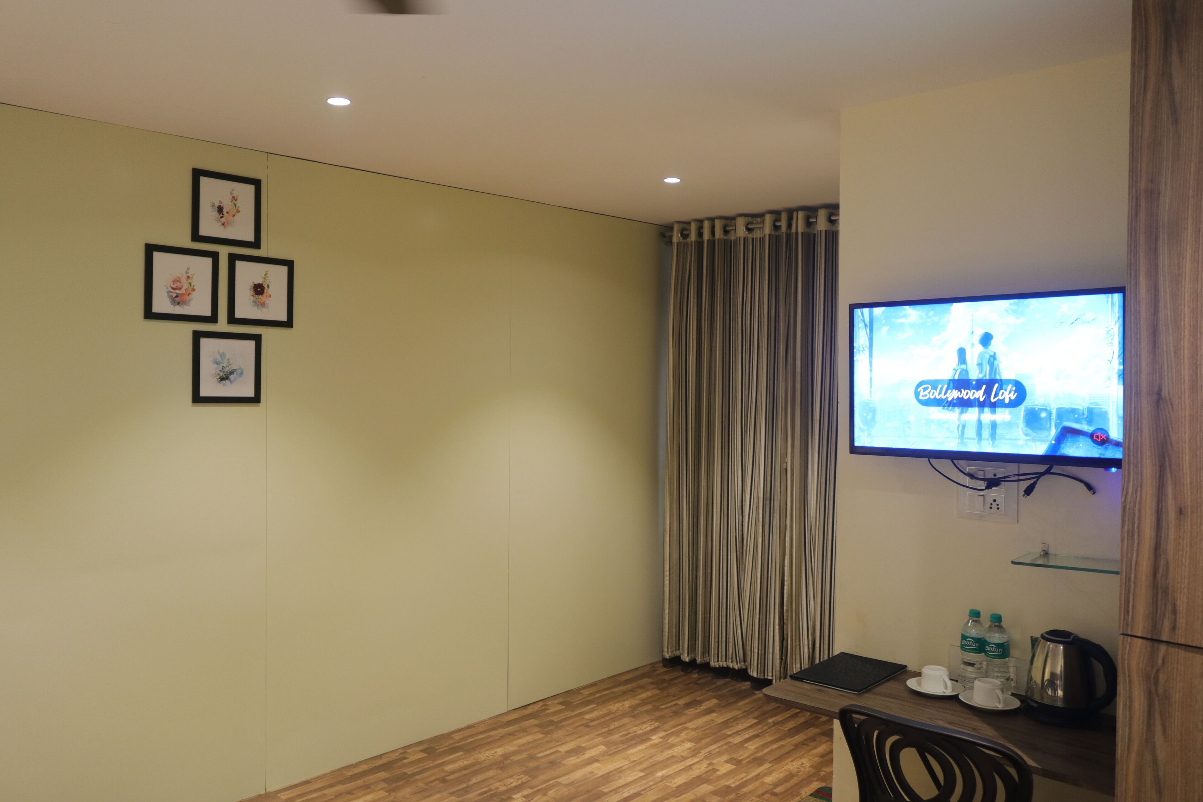 A hotel room with a wooden floor, a TV mounted on the wall, a desk with a laptop, a coffee maker, and a shelf with water bottles.