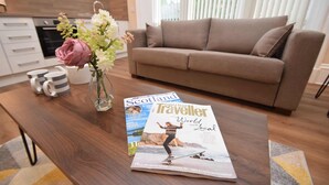 A modern living room with a brown sofa, a wooden coffee table with magazines, and a vase with flowers. - Rosedene Highland House - Emperor Apartment 1 (Inverness)