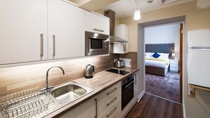 A compact kitchen with a sink, kettle, microwave, and toaster. A bed with a tufted headboard is visible through an open door. - Rosedene Highland House Emperor Studio Apart 3 (Inverness)