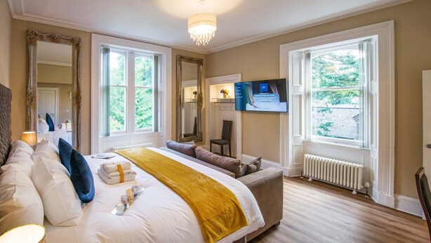 A spacious bedroom with a large bed, a television, and a view of the outdoors through the windows. - Rosedene Highland House - Emperor Apartment 6 (Inverness)