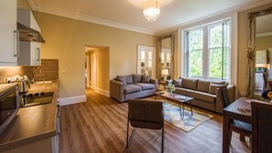 A spacious living room with a sofa, coffee table, and a dining area with a table and chairs. There is a kitchen with a stove and cabinets. - Rosedene Highland House - Emperor Apartment 7 (Inverness)