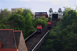 Una vía de tren con dos trenes rojos ascendiendo una colina, rodeada de exuberante vegetación y edificios con estilos arquitectónicos variados.
