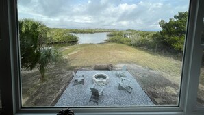 Vista a través de una ventana de un patio trasero con una chimenea, sillas y un lago.