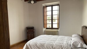Un dormitorio con piso de madera, cama con ropa blanca, ventana con contraventanas de madera y un radiador.